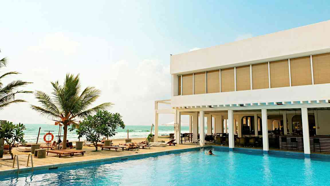 En udendørs pool med liggestole og palmer ved siden af et moderne hotel med udsigt over havet. (AI-genereret tekst)