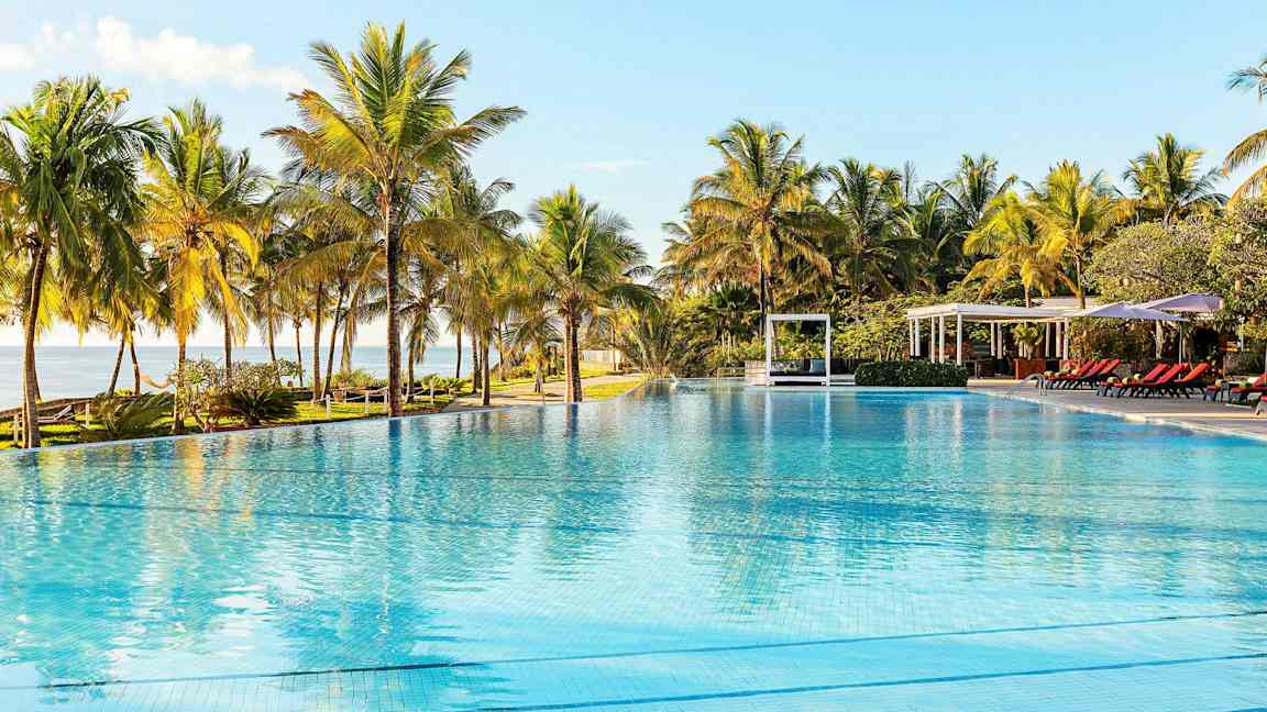 En stor udendørs pool omgivet af palmer, med liggestole og pavilloner i baggrunden. (AI-genereret tekst)