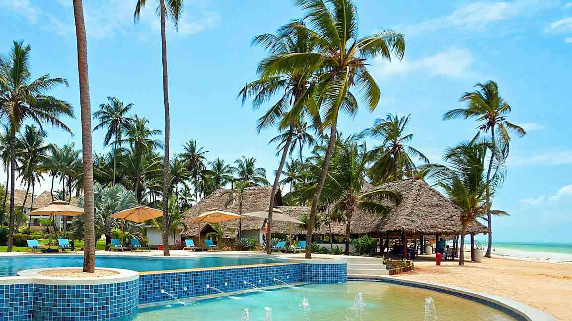 Et poolområde nær stranden omgivet af palmer og parasoller. (AI-genereret tekst)