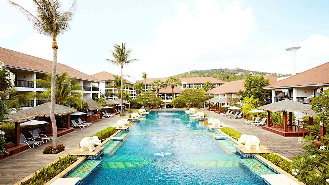 En stor pool på et feriested omgivet af palmer, liggestole og bungalowlignende bygninger. (AI-genereret tekst)