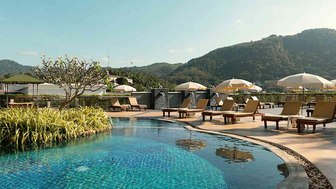 Udendørs pool med liggestole og parasoller, omgivet af grønne områder og bjerge i baggrunden. (AI-genereret tekst)