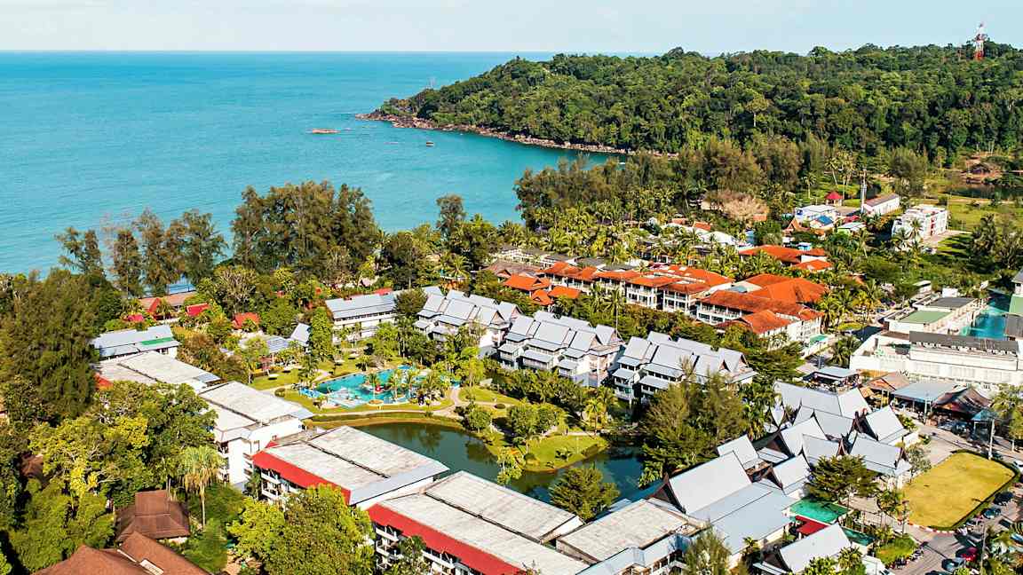 Luftfoto af et hotelkompleks omgivet af grønt og beliggende ved havet. (AI-genereret tekst)