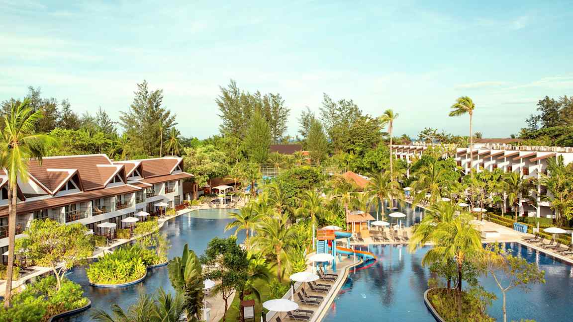 Hotelområde med en stor pool omgivet af palmer og bygninger. (AI-genereret tekst)