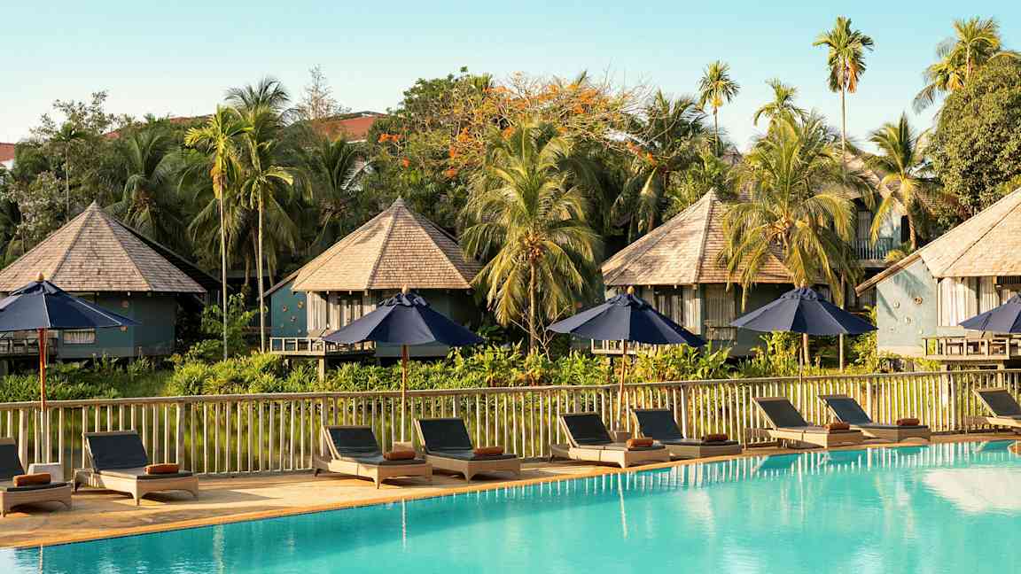 Liggestole og parasoller ved en swimmingpool med bungalowlignende bygninger og vegetation i baggrunden. (AI-genereret tekst)