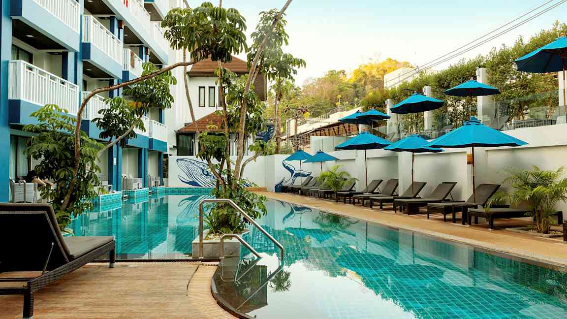 En udendørs swimmingpool med liggestole og parasoller, omgivet af vegetation og en fler-etagers hotelbygning. (AI-genereret tekst)