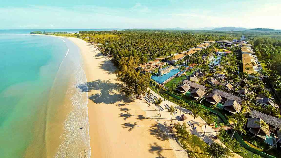 Luftfoto af en lang sandstrand med turkis vand og et feriekompleks med bungalower og pools. (AI-genereret tekst)