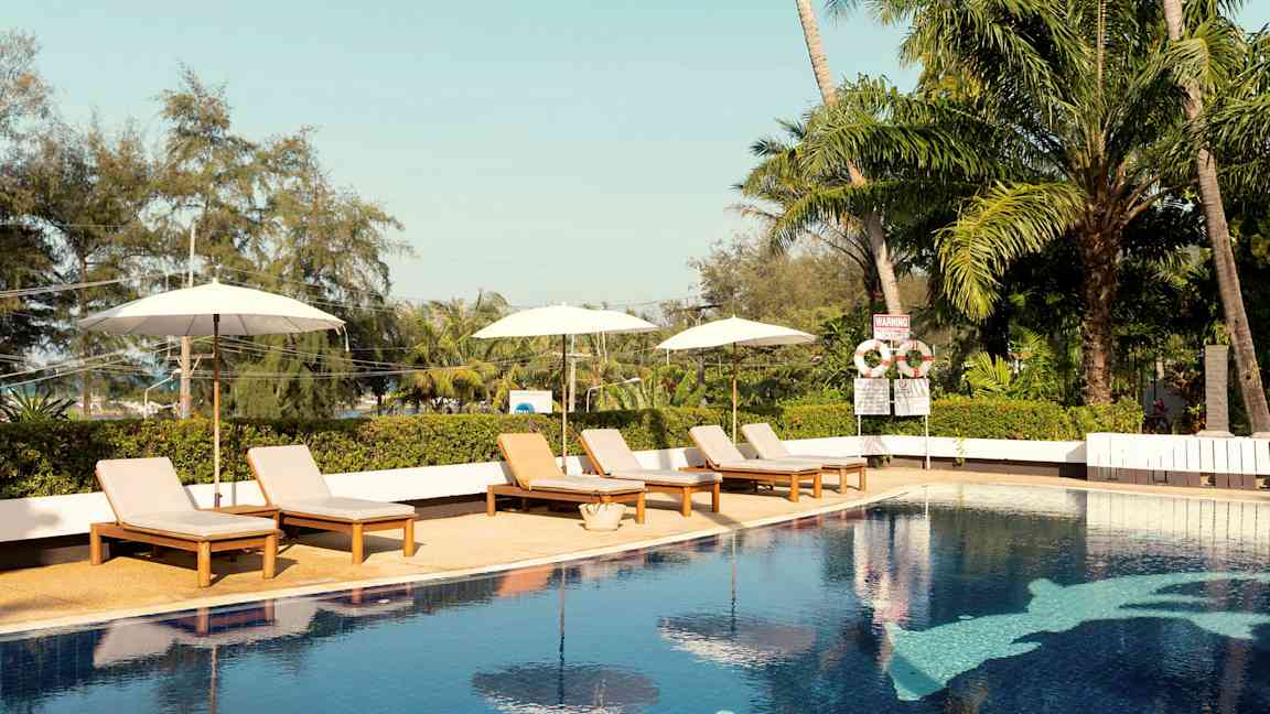 En udendørs pool med solsenge og parasoller. Tropiske træer i baggrunden. (AI-genereret tekst)