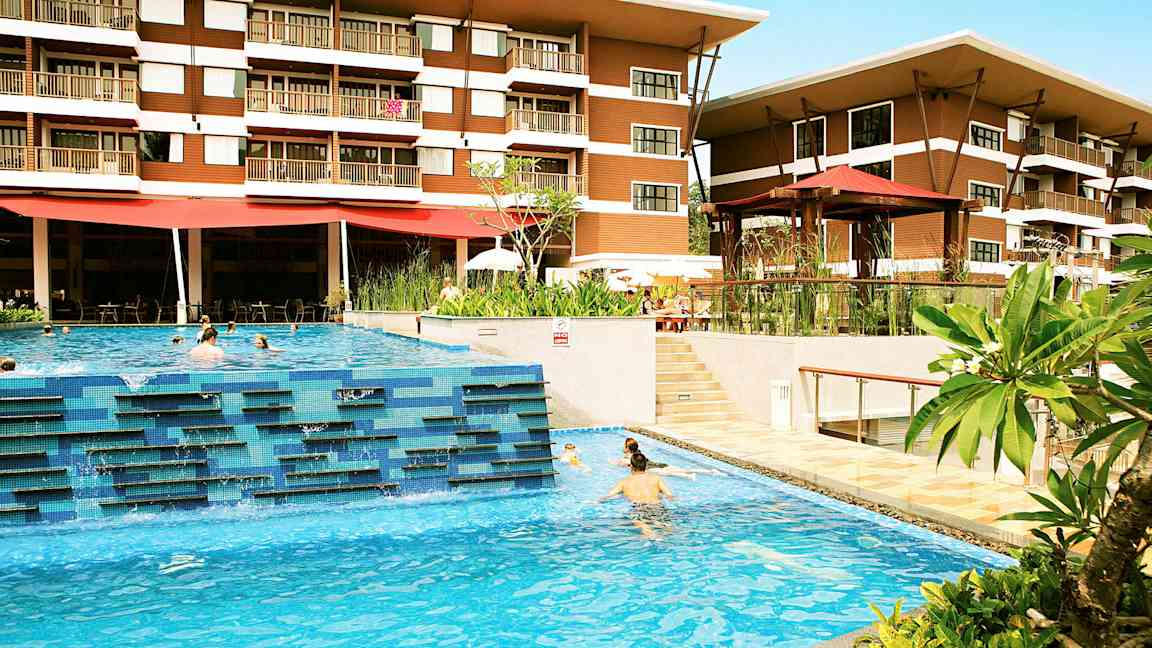 Et hotel med to etager er synligt bag en udendørs pool, hvor mennesker svømmer. Grønne planter og terrasseområder omkranser poolområdet. (AI-genereret tekst)