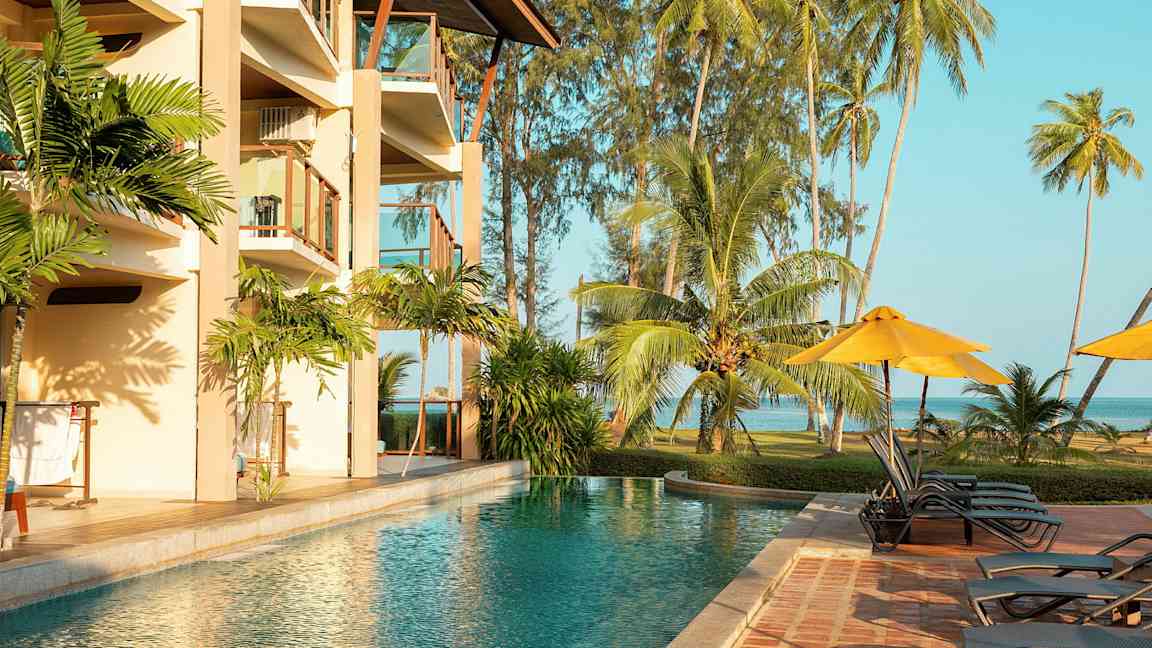 En udendørs pool ved siden af et hotel med altaner, omgivet af tropiske palmer og græsareal ved havet. (AI-genereret tekst)