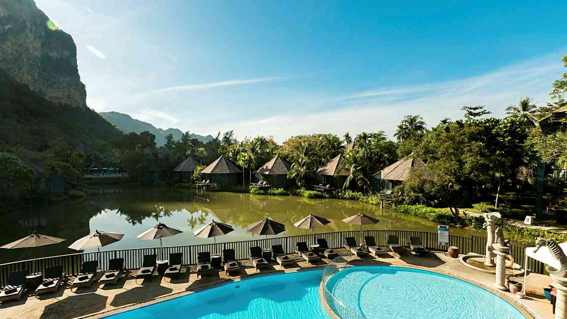 Et idyllisk ferieanlæg med bungalower ved en fredelig dam og en pool med solsenge i forgrunden. (AI-genereret tekst)