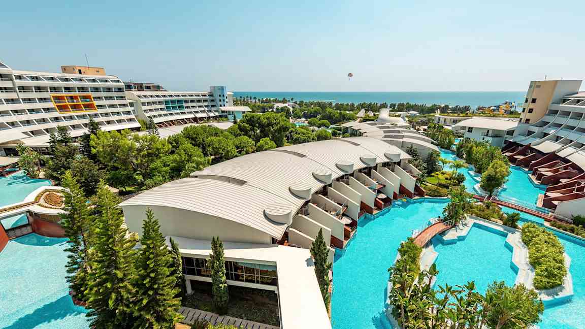 Udsigt over et hotelområde med moderne bygninger, store swimmingpools og frodige haver, der strækker sig mod havet. (AI-genereret tekst)