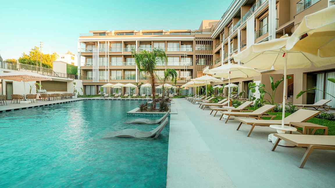 En hotelpool med solsenge og parasoller ved siden af moderne bygninger. (AI-genereret tekst)