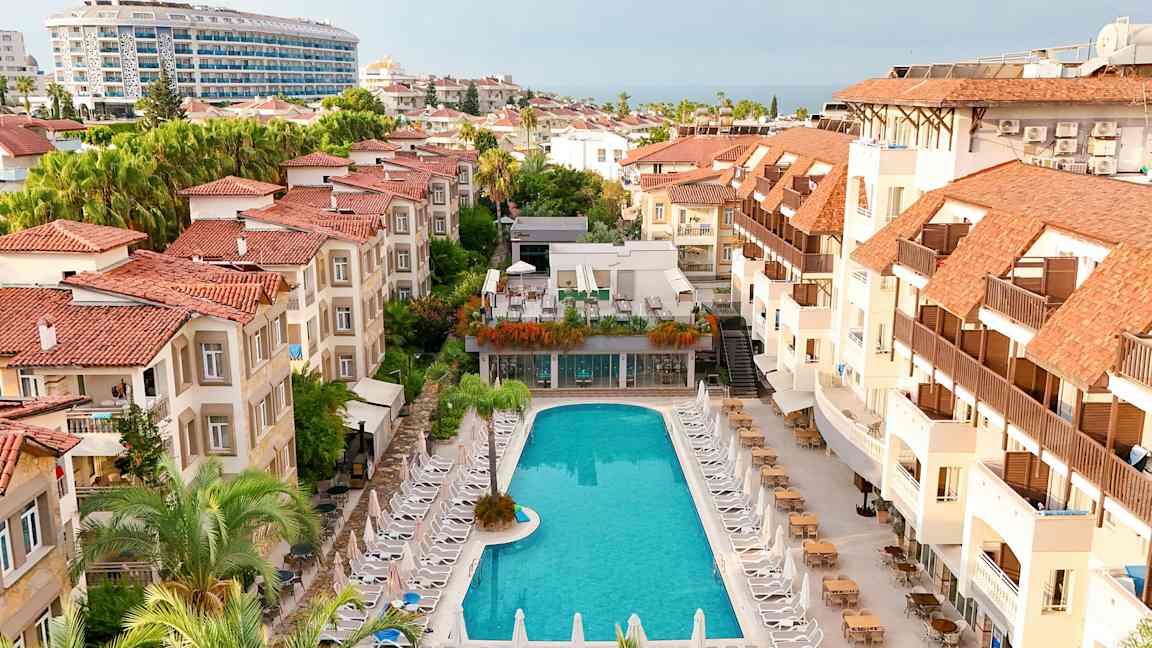 Hotelområde med en pool omgivet af liggestole og bygninger med røde tage. (AI-genereret tekst)