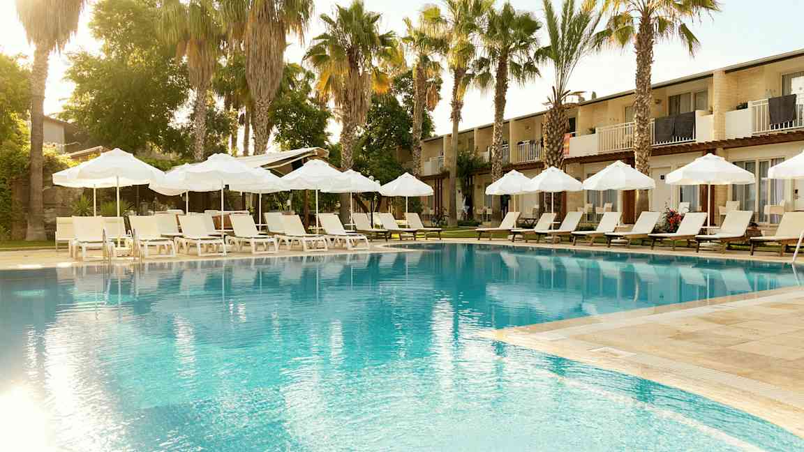 Pool med liggestole og parasoller, omgivet af palmer og bygninger i baggrunden. (AI-genereret tekst)