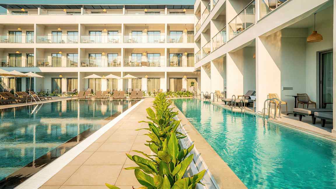 Billede af hotel med pool foran bygningen med liggestole og grønne planter. (AI-genereret tekst)