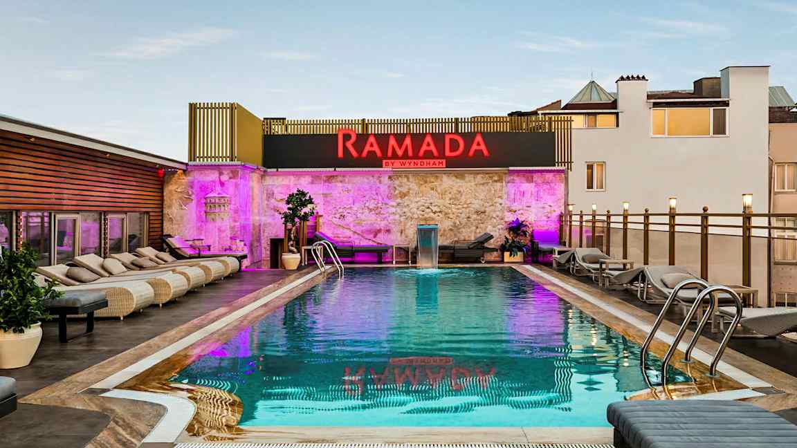 Billede af en udendørs pool med liggestole omkring og et skilt med teksten Ramada by Wyndham. (AI-genereret tekst)
