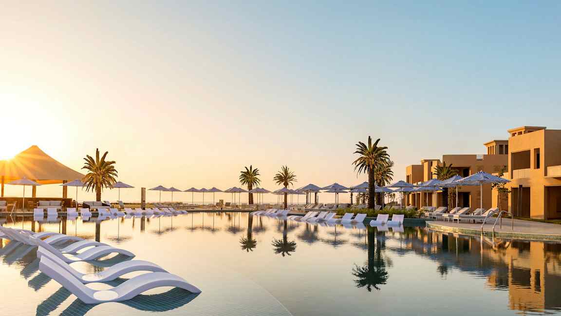 En udsigt over en infinitypool med liggestole og parasoller, omgivet af palmer og ferieboliger ved solnedgang. (AI-genereret tekst)