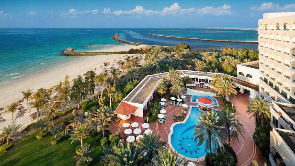 Et billede af et hotel ved stranden med en udendørs pool, liggestole og omkringliggende beplantning. (AI-genereret tekst)