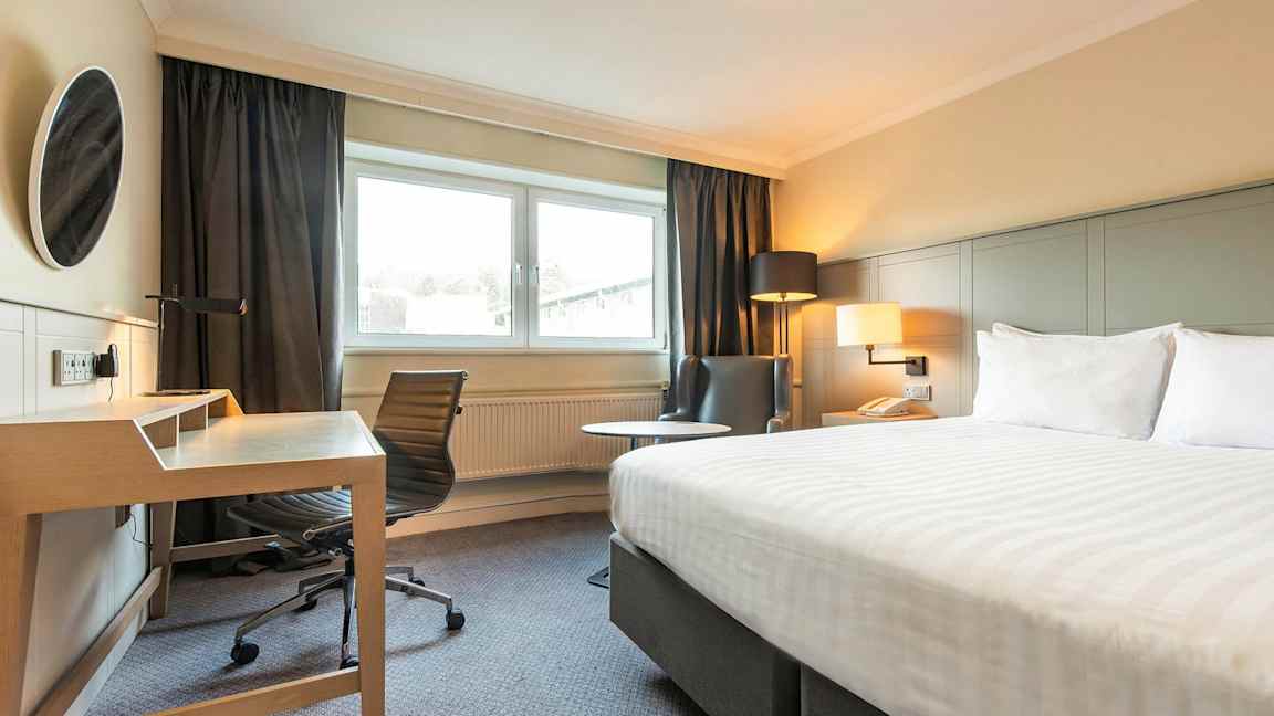 Billede af et hotelværelse med en seng, skrivebord og stol, samt en lænestol ved siden af et vindue. (AI-genereret tekst)