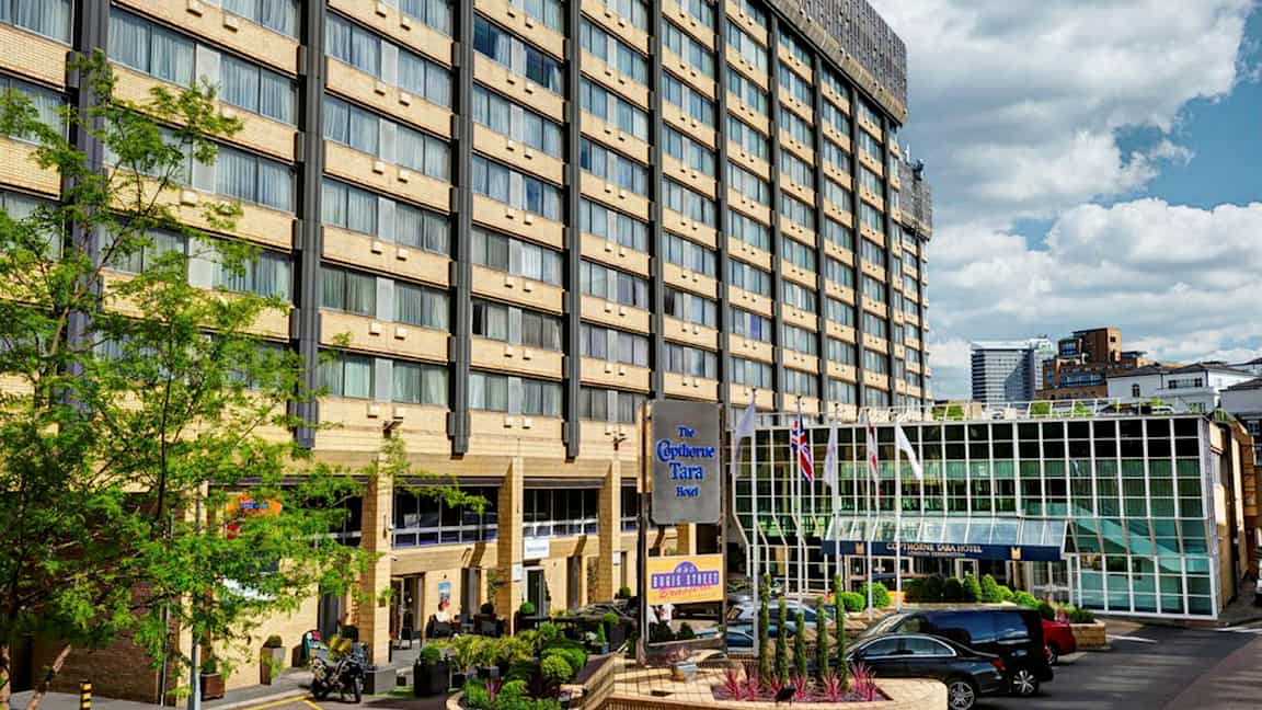 Hotelbygning med flere etager, en glasindgang og en parkeringsplads foran. (AI-genereret tekst)