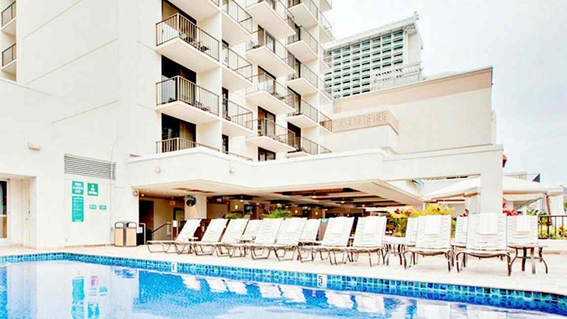 Billede af en bygning med altaner, en pool og liggestole. (AI-genereret tekst)