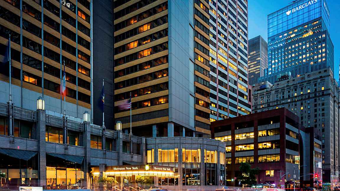 Et billede af et bymiljø med høje bygninger, hvoraf en bygning har teksten "Sheraton New York" ved indgangen. Taxaer er synlige på gaden. (AI-genereret tekst)