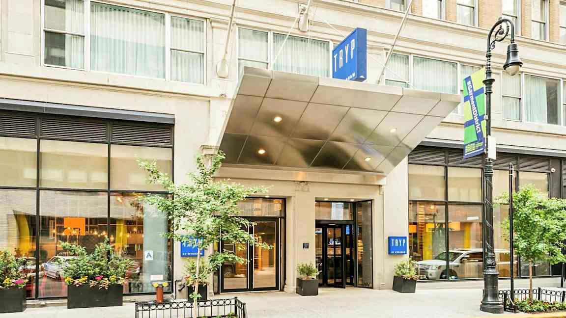 Indgangen til et hotel med skiltet "TRYP". Bygningen har store vinduer og plantekasser. (AI-genereret tekst)