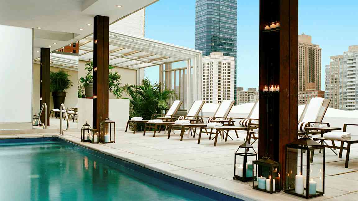 En moderne udendørs pool med solsenge og tændte lygter i et bymiljø. (AI-genereret tekst)