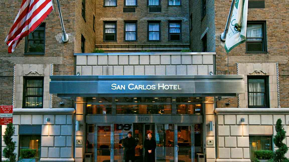 Indgangen til San Carlos Hotel, en bygning facade med mursten og flag. (AI-genereret tekst)