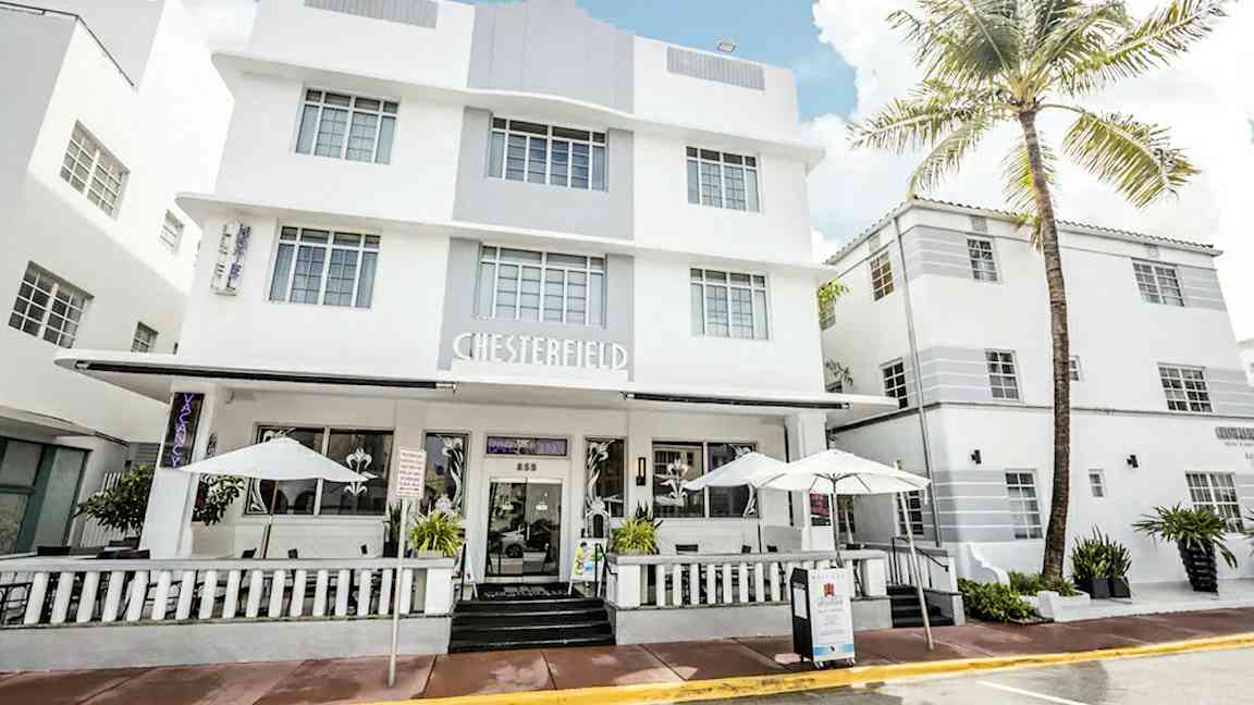 Et hvidt hotel i moderne arkitekturstil med store vinduer og en veranda med parasoller. (AI-genereret tekst)