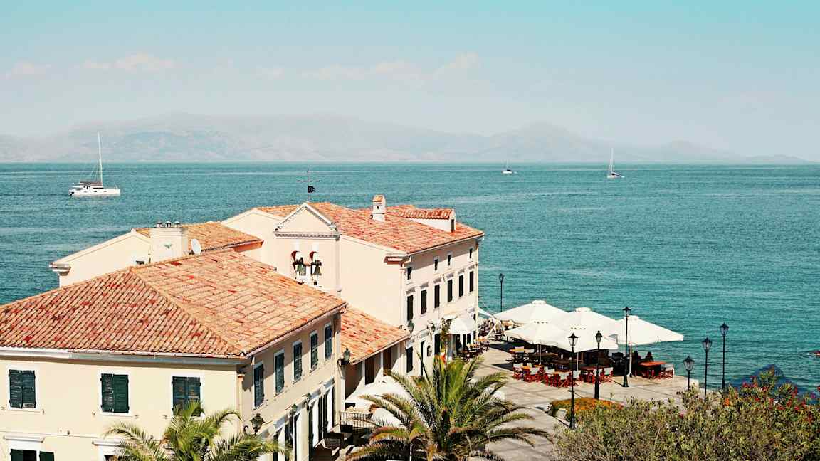 Billede af bygninger med tegltage ved havet, palmer og parasoller er synlige. (AI-genereret tekst)