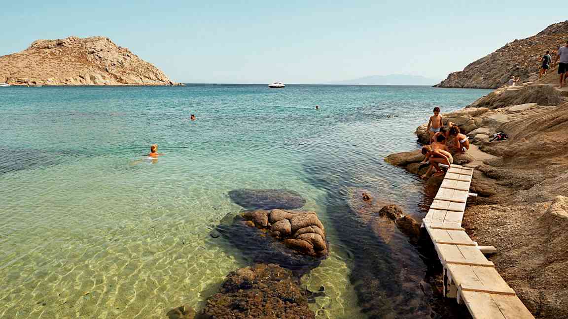 Personer ved en klippe nær en klarblå havbugt med badende. (AI-genereret tekst)