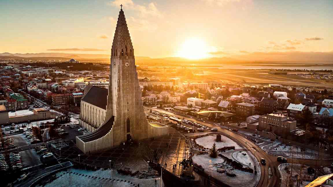 En kirke med moderne arkitektur og en høj tårnstruktur, omgivet af en by og et landskab under en solnedgang. (AI-genereret tekst)