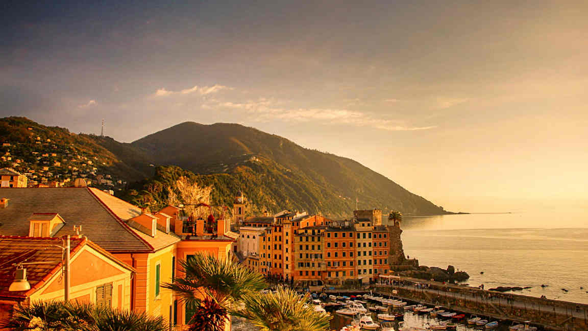 Billede af en kystby ved havet med små både i havnen og bjerge i baggrunden. (AI-genereret tekst)