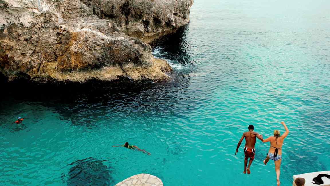 To personer hopper fra en klippe ned i blåt vand. (AI-genereret tekst)