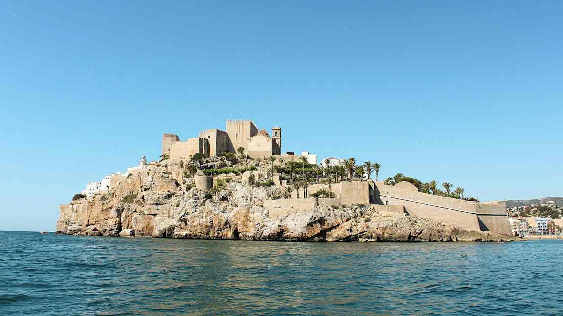 En borg og bygninger på en klippe ved havet, omgivet af palmer. (AI-genereret tekst)