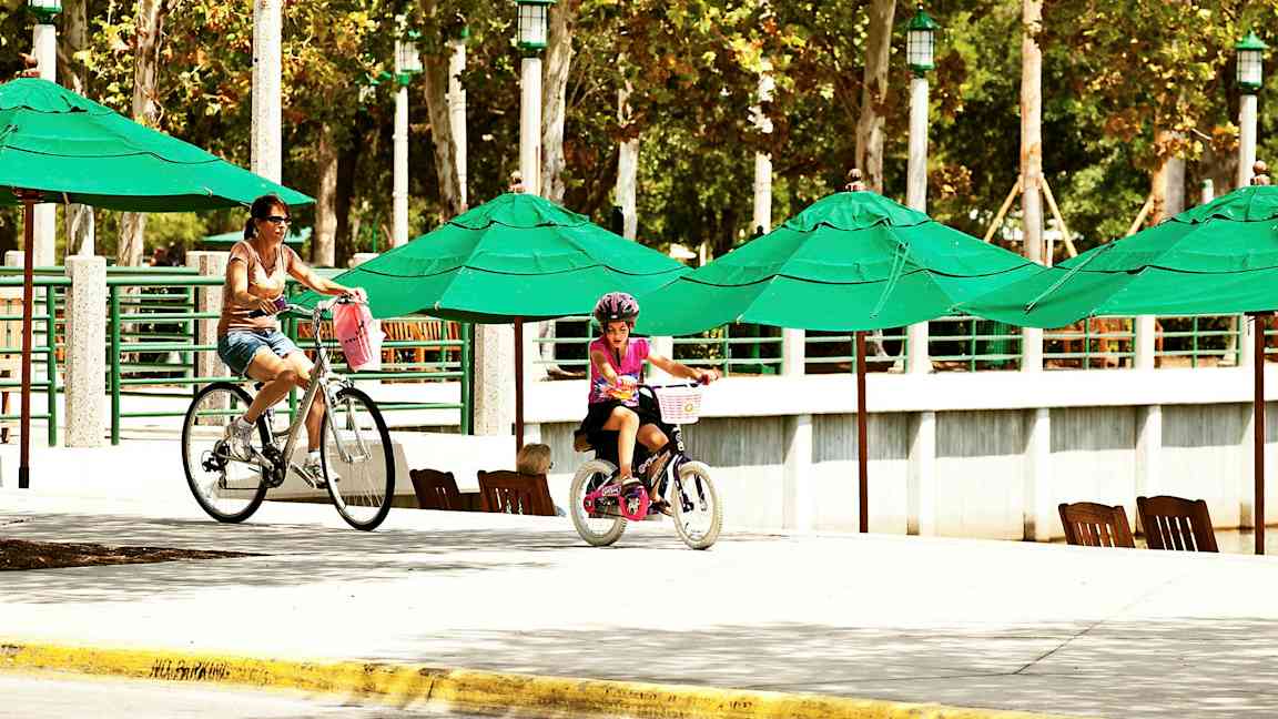 Personer cykler ved et område med grønne parasoller og træer i baggrunden. (AI-genereret tekst)