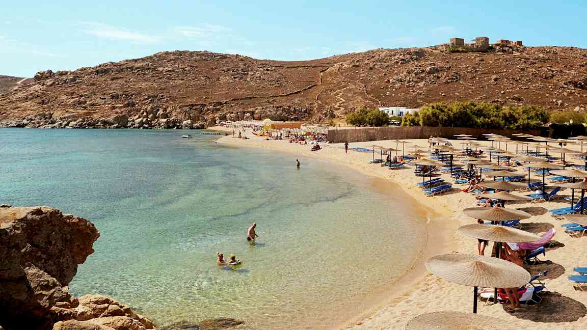 En strand med parasoller og liggestole, folk svømmer i det klare blå vand. (AI-genereret tekst)