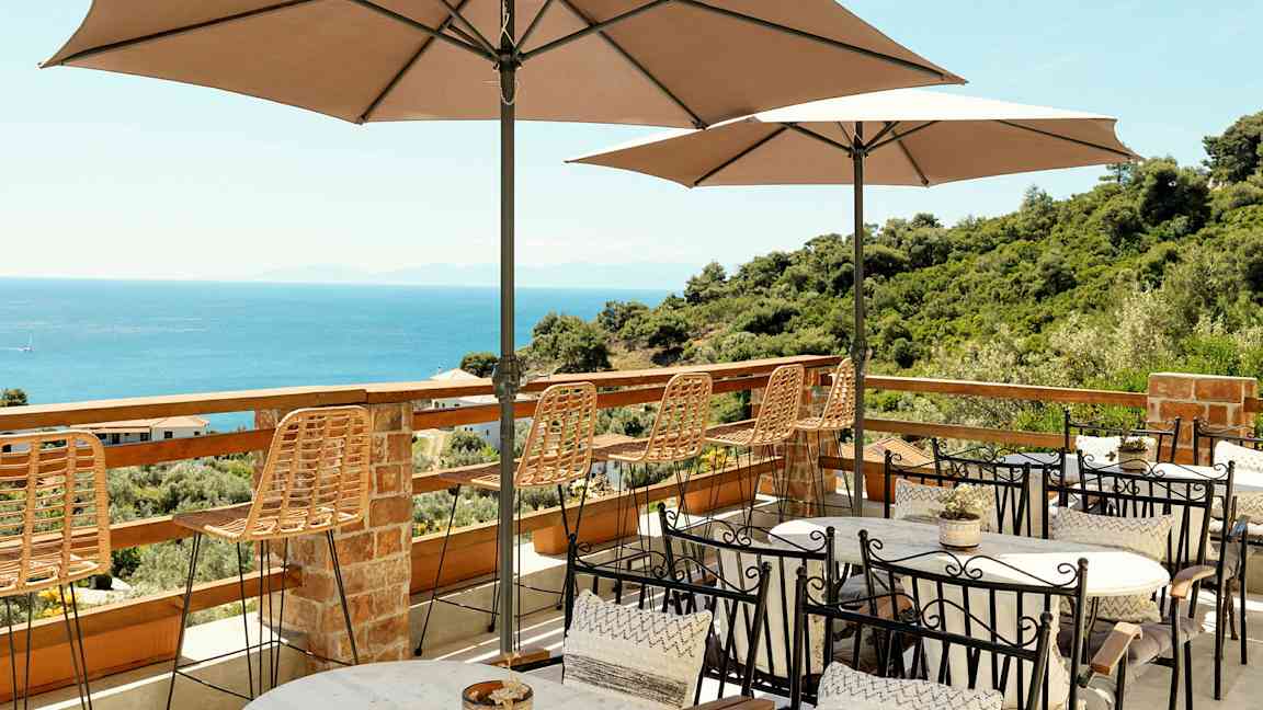 Terrasse med borde, stole og parasoller med udsigt over havet og frodige omgivelser. (AI-genereret tekst)