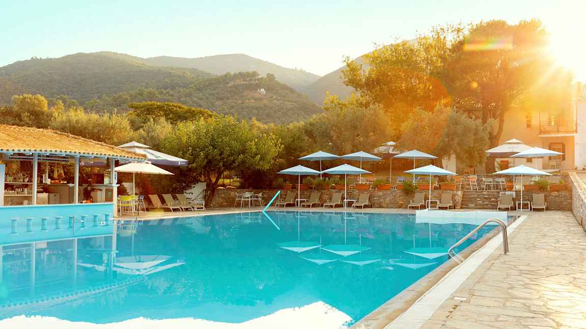 En udendørs pool med liggestole og parasoller, omgivet af træer og bygninger i et bjergrigt landskab. (AI-genereret tekst)