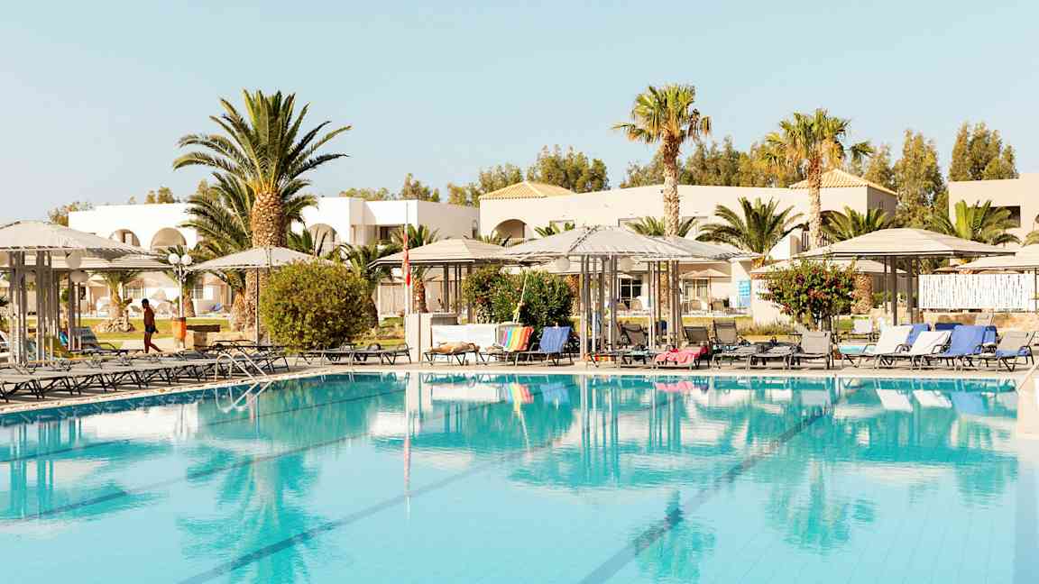 Billede af en pool med solsenge og parasoller, omkranset af træer og bygninger. (AI-genereret tekst)