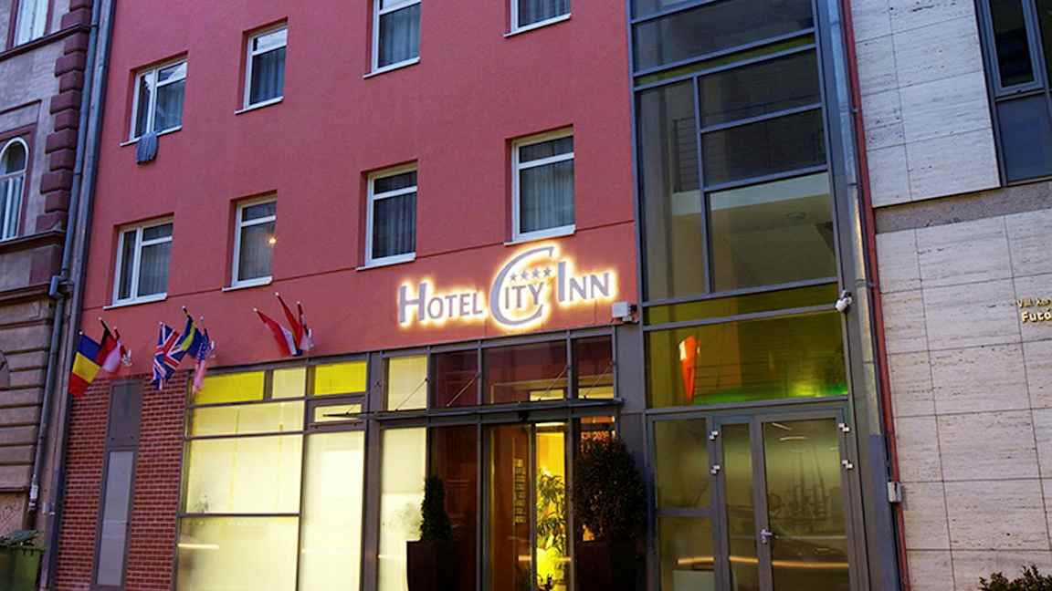 Uden for en hotelbygning med et skilt, der lyder "Hotel City Inn", og flere flag ved indgangen. (AI-genereret tekst)