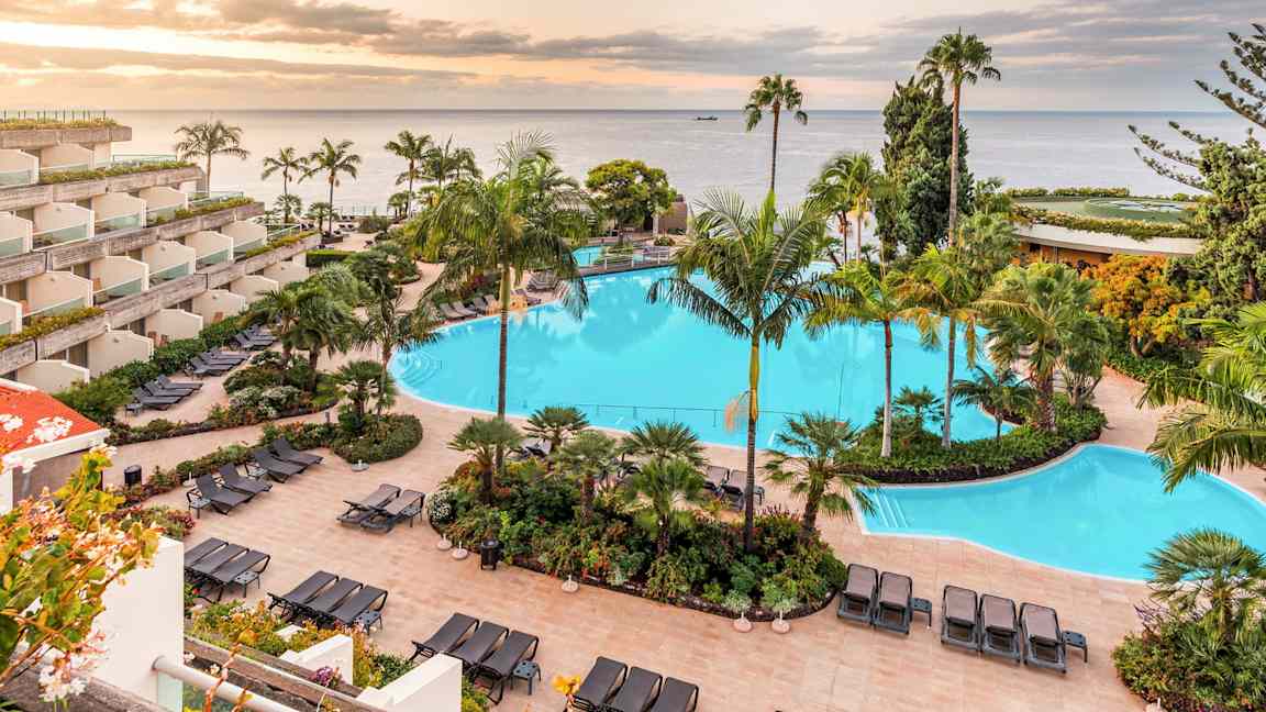 Billede af et hotelområde med en stor swimmingpool omgivet af liggestole og tropiske planter. Havet og en solnedgang ses i baggrunden. (AI-genereret tekst)