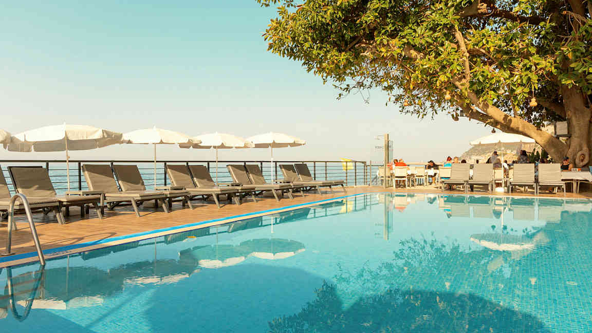 En udendørs pool med liggestole og parasoller ved siden af samt et træ og en træterrasse med siddepladser. (AI-genereret tekst)