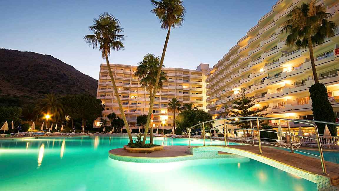 Et billede af et hotelområde ved skumring, med en oplyst pool, en bro og høje bygninger i baggrunden. (AI-genereret tekst)