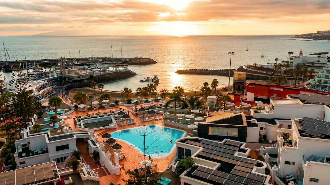 Billede af et kystområde med poolområde og bygninger i forgrunden, en havn med både og solnedgang over havet. (AI-genereret tekst)