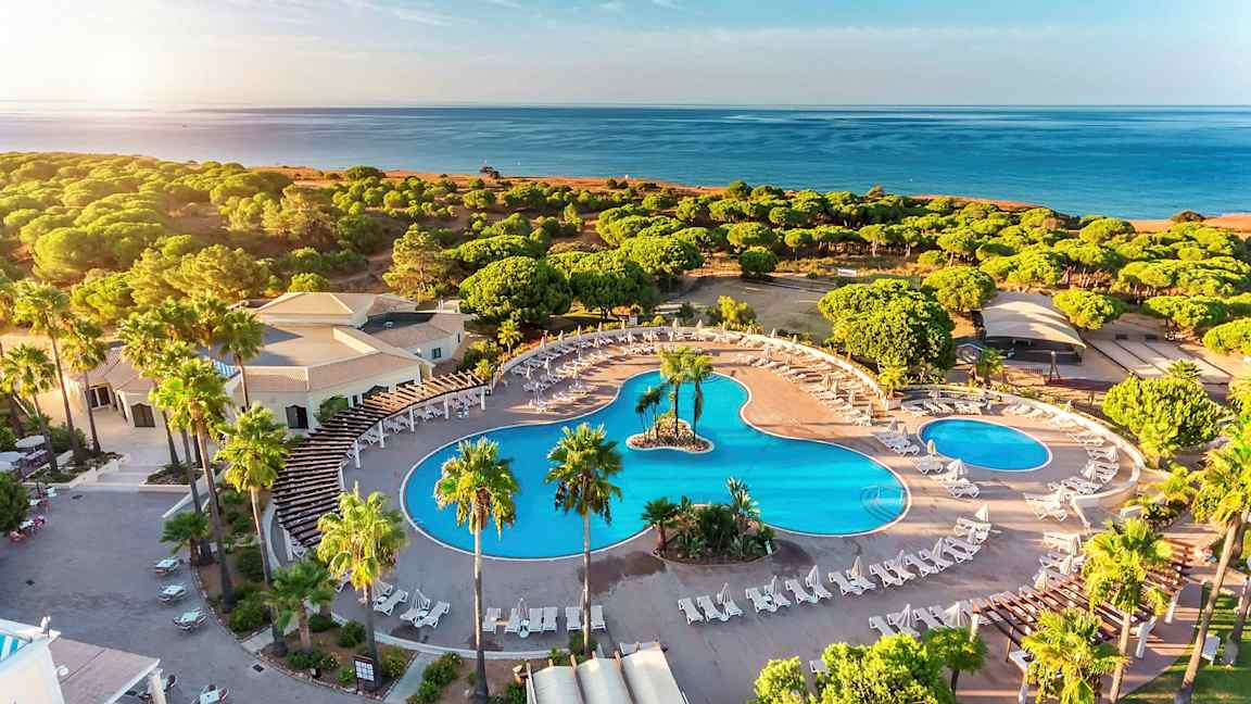 Luftfoto af et hotelområde med store swimmingpools, liggestole og omgivelser med grønne områder nær stranden. (AI-genereret tekst)