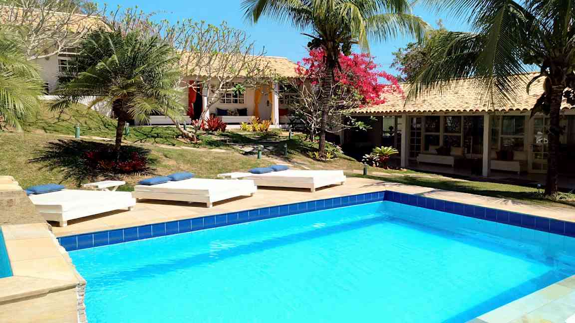 En pool omgivet af solsenge og tropiske planter ved en bygning med terracottatag. (AI-genereret tekst)