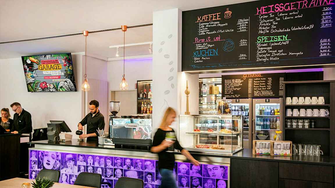 En caféindretning med en disk med en kaffemaskine, bagværk i en glasmontre og en menu på væggen ovenfor. Personale står bag disken. (AI-genereret tekst)
