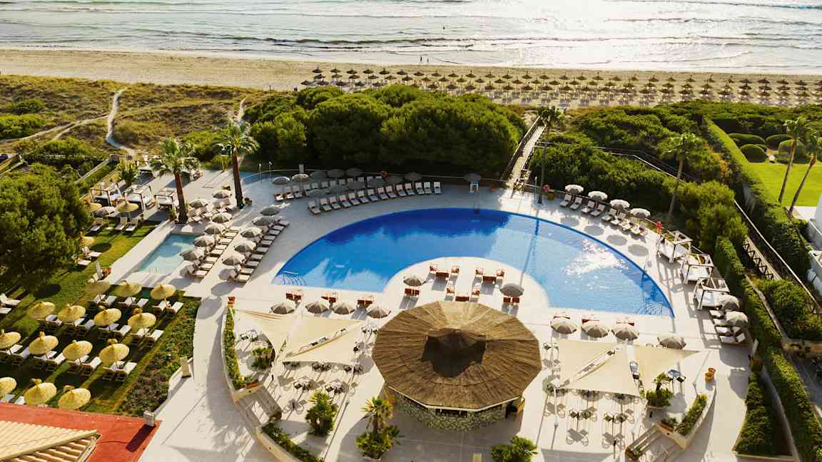 Udsigt over et poolområde med liggestole og parasoller, omgivet af grønt og tæt på stranden. (AI-genereret tekst)